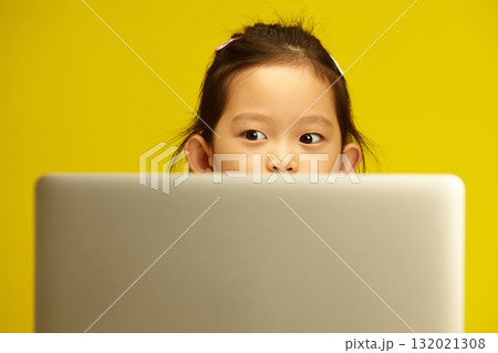 Pensive Asian Preschool Girl with a big opened eyes Engaged in Distance Learning via Laptop sitting against yellow isolated background Pensive Asian Preschool Girl with a big opened eyes Engaged in Distance Learning via Laptop sitting against yellow isolated background 132021308
