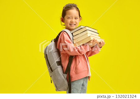 Cheerful little korean first grader girl in casual clothes with many books for studying at school and backpack on shoulders isolated over yellow background 132021324