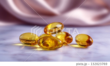 multiple golden, translucent soft gel capsules, likely containing omega-3 or other oil-based supplements, arranged on a light-colored surface with a blurred satin-like background 132023769