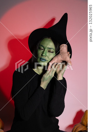A wicked witch in a black hat holds a mysterious object to her face on Halloween 132025965