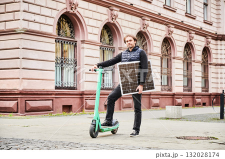 Man rides electric scooter while carrying solar panel. Integration of solar power as sustainable energy source for charging electric scooters, promoting eco-friendly urban transportation. Man rides electric scooter while carrying solar panel. Integration of solar power as sustainable energy source for charging electric scooters, promoting eco-friendly urban transportation. 132028174
