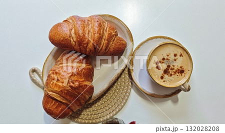 Two golden croissants served on a ceramic plate with a cup of cappuccino sprinkled with cinnamon. Concept of cozy breakfast, comfort, and slow morning indulgence at home or cafe 132028208