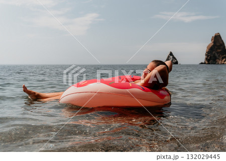 Woman, Inflatable Ring, Beach - Relaxing on a pink floatie in the sea by a rocky shore. Woman, Inflatable Ring, Beach - Relaxing on a pink floatie in the sea by a rocky shore. 132029445