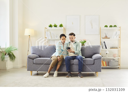 Happy couple sitting on sofa in living room at home and using smartphone together browsing apps. Happy couple sitting on sofa in living room at home and using smartphone together browsing apps. 132031109