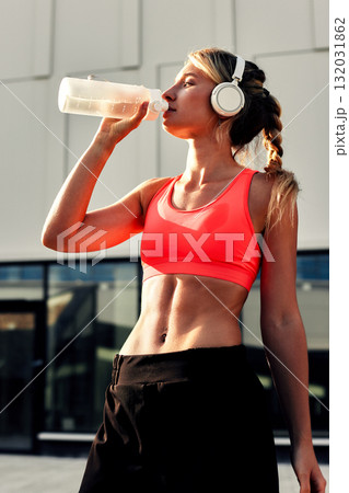Fit woman in bright sports bra drinking from shaker bottle while wearing headphones outdoors 132031862