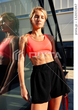 Athletic woman in coral sports bra and black shorts leaning by glass building during outdoor workout Athletic woman in coral sports bra and black shorts leaning by glass building during outdoor workout 132031947
