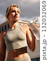 Focused athlete woman drinks water after outdoor workout, wearing a gray sports bra on a sunny day 132032069