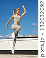 Athletic woman in gray workout set performs a dynamic jump on rooftop under a clear blue sky 132032092