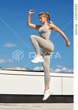 Athlete woman mid-air jump in athletic wear during outdoor rooftop workout against blue sky 132032093