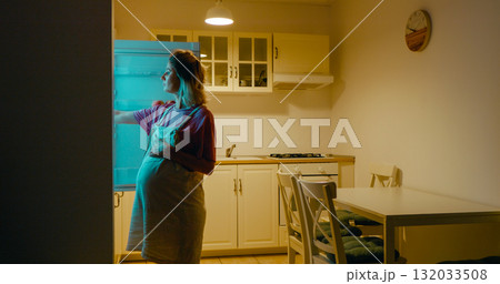 Pregnant woman opening refrigerator and eating cake at home. 132033508