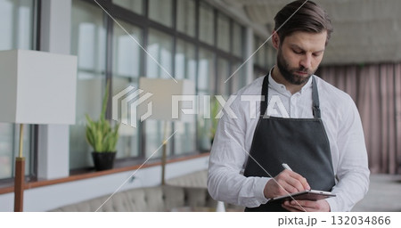 Young waiter man writes down an order in a notebook in a restaurant. Portrait of a young waiter in a white shirt and apron smiling in a restaurant. Young waiter man writes down an order in a notebook in a restaurant. Portrait of a young waiter in a white shirt and apron smiling in a restaurant. 132034866