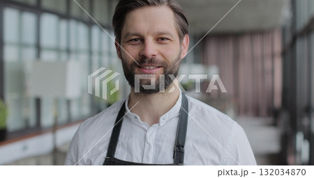Close up male restaurant waiter in apron standing in empty cafe hall, looking and smiling on camera. Close up of attractive man working in restaurant. 132034870