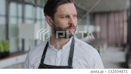 Close up male restaurant waiter in apron standing in empty cafe hall, looking and smiling on camera. Close up of attractive man working in restaurant. Close up male restaurant waiter in apron standing in empty cafe hall, looking and smiling on camera. Close up of attractive man working in restaurant. 132034871