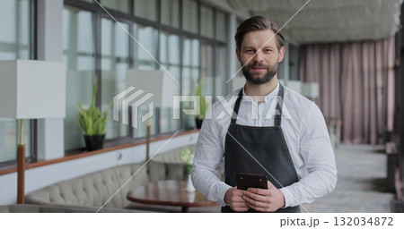 Portrait of a young waiter in a white shirt and apron smiling in a restaurant. Handsome guy expressing positivity while standing against the background of a restaurant. Male waiter in a cafeteria. 132034872