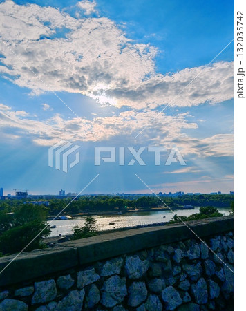 Scenic river view with sun rays through clouds. Panoramic view over a river and distant city skyline with dramatic sun rays breaking through the clouds above a stone wall. Scenic river view with sun rays through clouds. Panoramic view over a river and distant city skyline with dramatic sun rays breaking through the clouds above a stone wall. 132035742