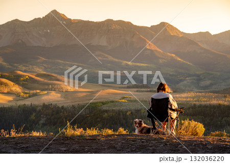 Female traveler sitting on a camping chair with her dog watching the mountain view during sunset. 132036220