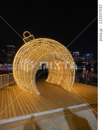 A glowing ornament-shaped tunnel sparkles with golden lights beside a calm lake. The city skyline shines in the background creating a festive night scene. 132037032