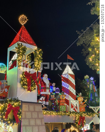 Santa Claus stands between glowing red towers surrounded by Christmas decorations and lights. The festive outdoor installation creates a joyful holiday atmosphere. Santa Claus stands between glowing red towers surrounded by Christmas decorations and lights. The festive outdoor installation creates a joyful holiday atmosphere. 132037118