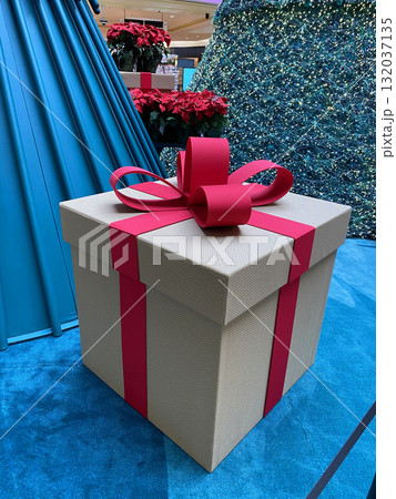 Close-up of a big decorative gift box with a red ribbon and bow inside a Christmas mall installation. The festive present stands near sparkling trees and bright holiday decor. 132037135
