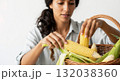 Woman peeling fresh corn on the cob from a wicker basket. Close-up of hands preparing a raw vegetable. Healthy eating and farm-to-table harvest concept Woman peeling fresh corn on the cob from a wicker basket. Close-up of hands preparing a raw vegetable. Healthy eating and farm-to-table harvest concept 132038360
