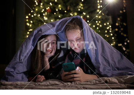 Two girls under a blanket looking at a smartphone near a Christmas tree with festive lights 132039048