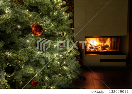 Beautiful close-up of a decorated Christmas tree with glowing lights and red ornaments beside a cozy fireplace, symbolizing warmth and festive comfort. Beautiful close-up of a decorated Christmas tree with glowing lights and red ornaments beside a cozy fireplace, symbolizing warmth and festive comfort. 132039051