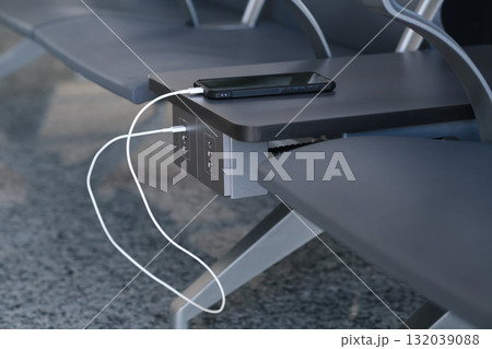 Charging station at an airport seating area with a smartphone plugged into an outlet during busy travel hours 132039088
