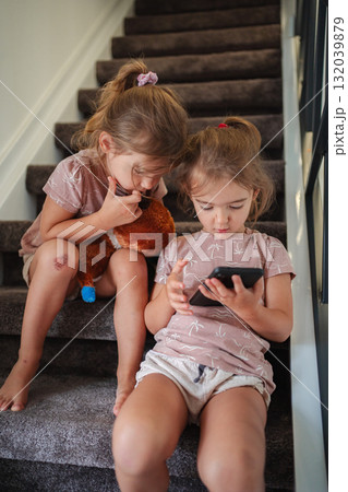 Two preschool girls playing with smartphone and teddy bear on stairs 132039879