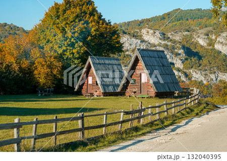 Closer view of two triangular wooden houses behind a rustic fence with mountains and trees in the background. Warm autumn scene, showcasing rural architecture and peaceful countryside atmosphere. 132040395