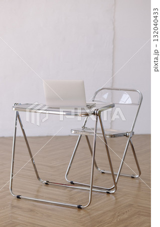 Minimalist chrome desk and transparent chair with laptop on wooden floor against white wall 132040433