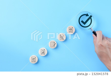 Document approval and quality assurance process concepts. Hand holding a magnifying glass focusing on check mark symbol with document icon, document approval, compliance, and quality assurance process 132041378