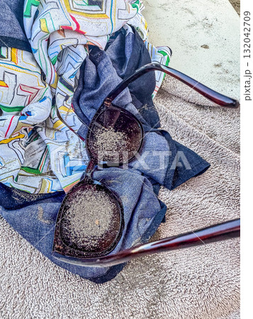 Close-up of fashionable sunglasses covered with sand resting on colorful fabric and a towel, symbolizing summer vacation, beach lifestyle, and trave. High quality photo 132042709