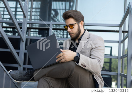 Man in suit and sunglasses sitting on steps, focused on laptop screen.  132043313