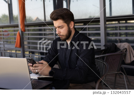 The man in the image is using his phone while sitting at a table with a laptop 132043557