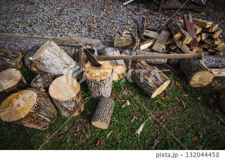 An axe stuck in a log surrounded by chopped firewood pieces and branches on the grass near a gravel area 132044458