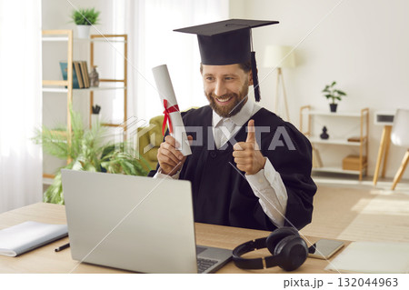 Man in graduate gown sitting at desk with diploma showing thumb up sign during online video call. Man in graduate gown sitting at desk with diploma showing thumb up sign during online video call. 132044963
