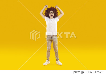 Funny boy in red curly wig screaming with joy with open mouth and with his hands up in fist. Funny boy in red curly wig screaming with joy with open mouth and with his hands up in fist. 132047570