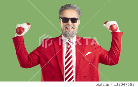 Funny modern Santa Claus in a stylish red suit and striped tie holds dumbbells showing how strong he is. 132047588