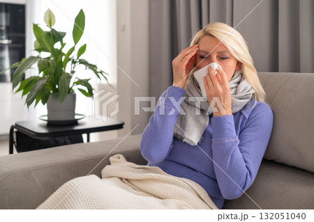 Sick middle aged woman sitting on sofa, wrapped in blanket, blowing her nose into a tissue while feeling headache. Concept of flu, cold, covid, and home rest. Sick middle aged woman sitting on sofa, wrapped in blanket, blowing her nose into a tissue while feeling headache. Concept of flu, cold, covid, and home rest. 132051040