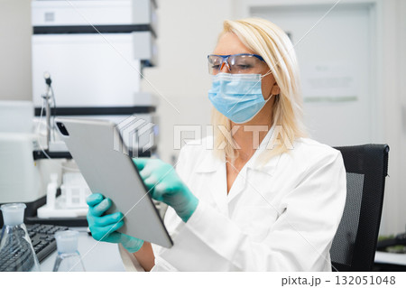 Female scientist in lab coat and mask using tablet to analyze data in laboratory. Female scientist in lab coat and mask using tablet to analyze data in laboratory. 132051048