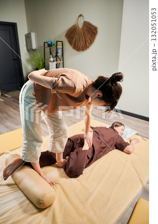 Masseuse making thai yoga massage. Woman therapist doing traditional massage treatment, stretching female patient's body. Client lying on mat, enjoying hand massage in calm atmosphere. Masseuse making thai yoga massage. Woman therapist doing traditional massage treatment, stretching female patient's body. Client lying on mat, enjoying hand massage in calm atmosphere. 132051053