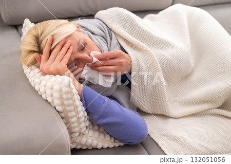 Middle aged blonde woman lying on couch at home, feeling sick and blowing her nose into a tissue. Concept of flu, cold, runny nose, and home rest during illness. Middle aged blonde woman lying on couch at home, feeling sick and blowing her nose into a tissue. Concept of flu, cold, runny nose, and home rest during illness. 132051056