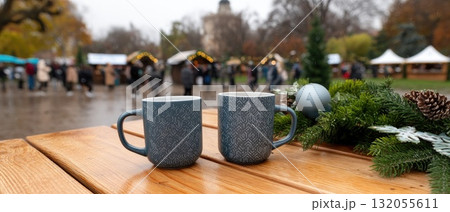 Two cups sit on a wooden table, surrounded by festive decorations and people enjoying a lively outdoor market on a rainy day Two cups sit on a wooden table, surrounded by festive decorations and people enjoying a lively outdoor market on a rainy day 132055611
