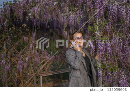 Woman Wisteria Phone Call Spring - A woman wearing a gray blazer stands in front of a flowering wisteria vine while on a phone call during springtime. Woman Wisteria Phone Call Spring - A woman wearing a gray blazer stands in front of a flowering wisteria vine while on a phone call during springtime. 132059608