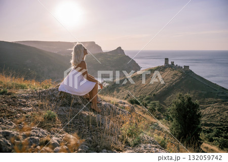 A woman is sitting on a hillside overlooking the ocean. She is wearing a white dress and has blonde hair. The scene is serene and peaceful, with the ocean in the background. A woman is sitting on a hillside overlooking the ocean. She is wearing a white dress and has blonde hair. The scene is serene and peaceful, with the ocean in the background. 132059742