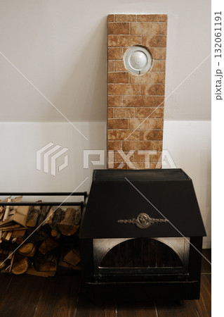 Black metal fireplace with stacked firewood and brick-patterned wall in a cozy home interior. Perfect for design, lifestyle, or winter comfort themes Black metal fireplace with stacked firewood and brick-patterned wall in a cozy home interior. Perfect for design, lifestyle, or winter comfort themes 132061191