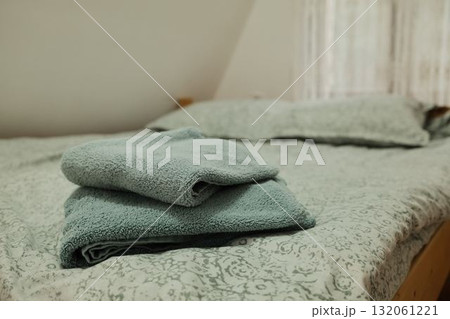 Folded soft towels placed on a neatly made bed with patterned linen and natural light through a lace curtain. Ideal for hospitality, home, or interior design use Folded soft towels placed on a neatly made bed with patterned linen and natural light through a lace curtain. Ideal for hospitality, home, or interior design use 132061221