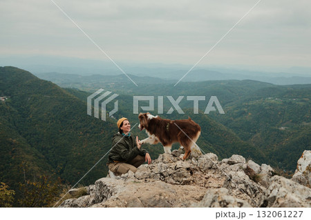 A smiling woman kneels on rocky cliffs as her dog Australian Shepherd gives her a paw with mountain landscapes in the background. Hiking with pets concept A smiling woman kneels on rocky cliffs as her dog Australian Shepherd gives her a paw with mountain landscapes in the background. Hiking with pets concept 132061227