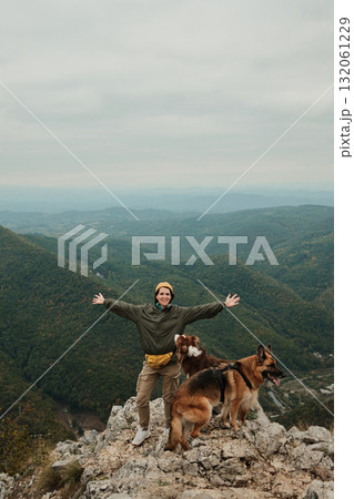 A happy woman raises her arms on the top of Mount Kablar, Serbia, with her two dogs Australian and German Shepherd beside her, enjoying the freedom of the mountains. Hiking with pets concept A happy woman raises her arms on the top of Mount Kablar, Serbia, with her two dogs Australian and German Shepherd beside her, enjoying the freedom of the mountains. Hiking with pets concept 132061229