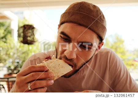 Man wearing brown beanie eats taco filled with vegetables at Mexican cafe table. Close front view portrait 132061457